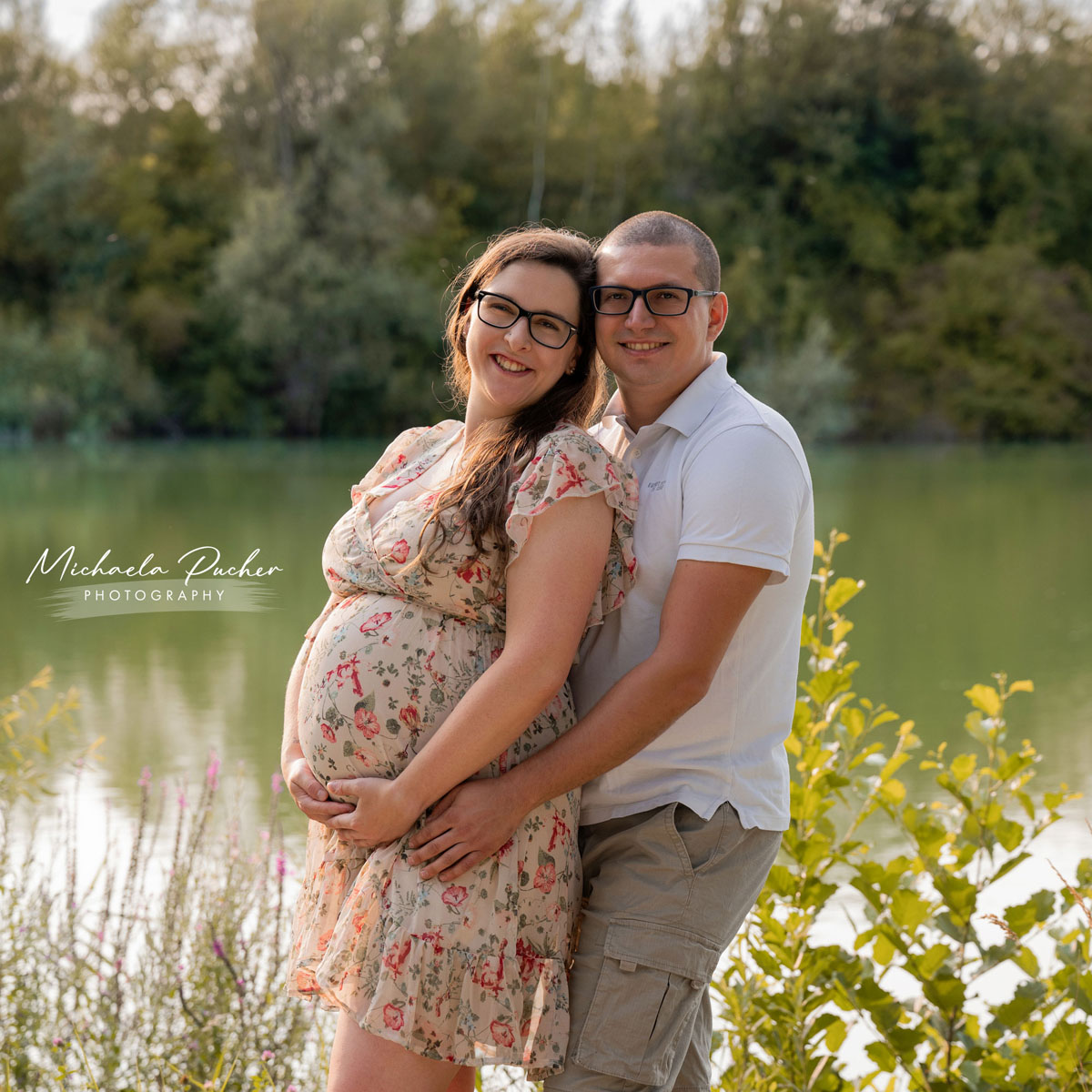 Schwangere Frau im geblümten Kleid steht am See und lächelt in die Kamera, während ihr Partner sie von hinten umarmt und ebenfalls lächelt.